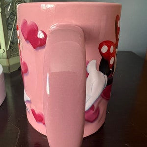 May include: Pink ceramic mug with a Minnie Mouse design featuring red polka dots and hearts. The mug has a large handle.