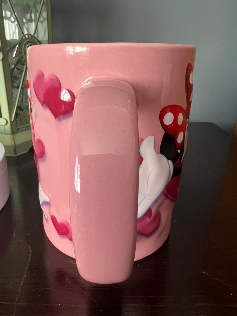 May include: Pink ceramic mug with a Minnie Mouse design featuring red polka dots and hearts. The mug has a large handle.