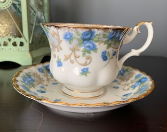 May include: A white teacup and saucer set with a blue and gold floral pattern. The cup has a delicate handle and a scalloped rim. The saucer has a matching floral design and a gold rim.