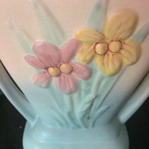 Puede incluir: Un jarrón de cerámica vintage con un esmalte azul claro y un diseño floral. El jarrón presenta dos flores, una rosa y una amarilla, con hojas verdes.