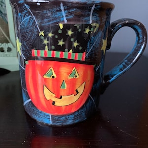 May include: A black ceramic mug with a blue and white spiderweb design. The mug features a red, orange, and yellow jack-o'-lantern face with a green nose and black eyes. The jack-o'-lantern is wearing a black and green hat with yellow stars.