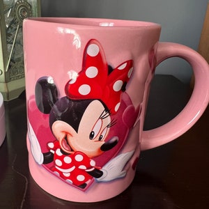 May include: Pink ceramic mug with a cartoon image of Minnie Mouse wearing a red bow with white polka dots. Minnie Mouse is smiling and holding a red heart with white polka dots. The mug has a white handle.