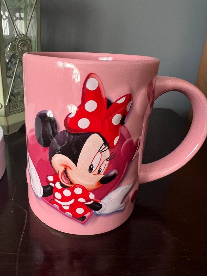 May include: Pink ceramic mug with a cartoon image of Minnie Mouse wearing a red bow with white polka dots. Minnie Mouse is smiling and holding a red heart with white polka dots. The mug has a white handle.