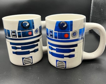 May include: Two white ceramic mugs with a blue and white R2-D2 design. The mugs have a handle on the right side.