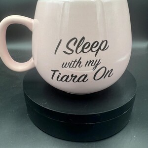 May include: A light pink ceramic mug with a rounded, bulbous shape and a curved handle. The front of the mug features the black printed text "I Sleep with my Tiara On" in a stylised script font. The interior of the mug is a pale blue colour.