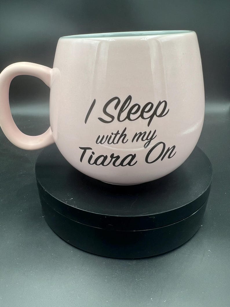 May include: A light pink ceramic mug with a rounded, bulbous shape and a curved handle. The front of the mug features the black printed text "I Sleep with my Tiara On" in a stylised script font. The interior of the mug is a pale blue colour.