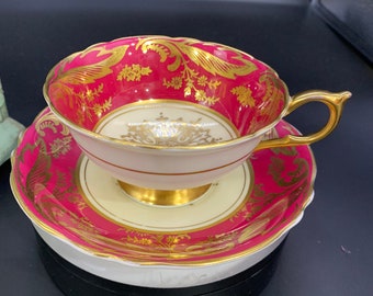 May include: A pink and gold teacup and saucer set with a floral design. The cup and saucer are both decorated with gold trim and a delicate floral pattern.
