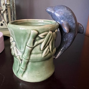 May include: A ceramic mug featuring a green bamboo design and a blue dolphin handle. The mug has a cylindrical shape with a wide opening and a glossy finish. The dolphin handle has a textured surface.