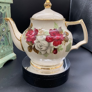 May include: A white teapot with a gold trim and a floral design featuring red and white roses. The teapot is sitting on a black pedestal.