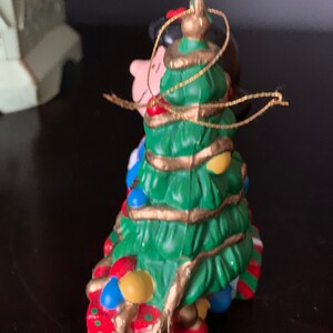 May include: A ceramic Christmas tree ornament featuring a cartoon couple standing in front of a decorated Christmas tree. The couple is holding presents and the tree is wrapped in gold ribbon.