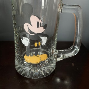 May include: A clear glass Mickey Mouse beer mug with a black and white image of Mickey Mouse on the front. Mickey Mouse is wearing yellow shoes.