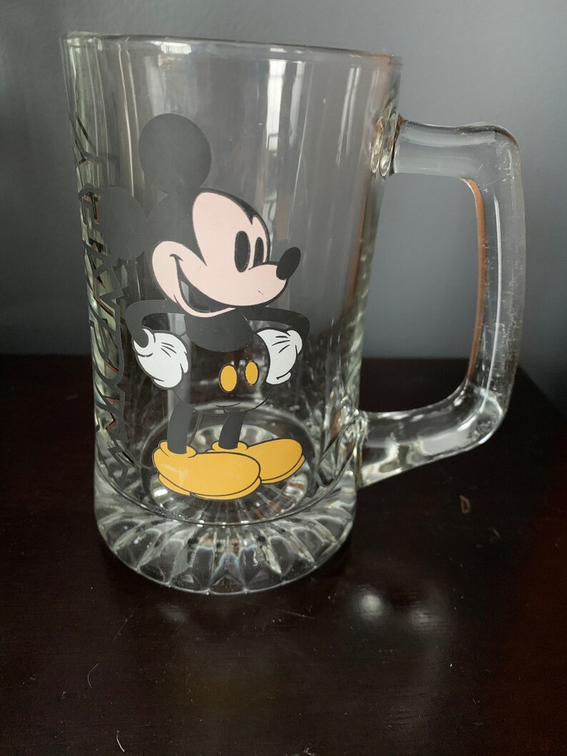 May include: A clear glass Mickey Mouse beer mug with a black and white image of Mickey Mouse on the front. Mickey Mouse is wearing yellow shoes.