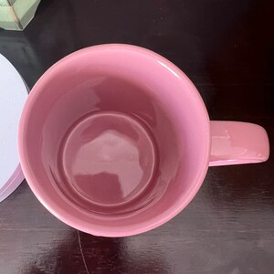 May include: A pink ceramic mug with a wide, round opening and a handle. The mug is empty and sitting on a dark brown wooden surface.