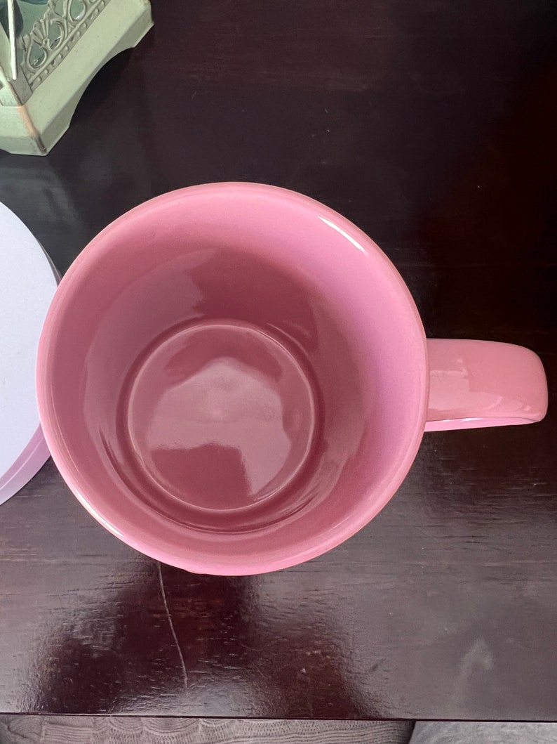 May include: A pink ceramic mug with a wide, round opening and a handle. The mug is empty and sitting on a dark brown wooden surface.