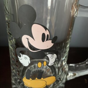 May include: A clear glass mug with a black and white image of Mickey Mouse on the front. Mickey Mouse is smiling and has his hands on his hips. He is wearing yellow shoes.