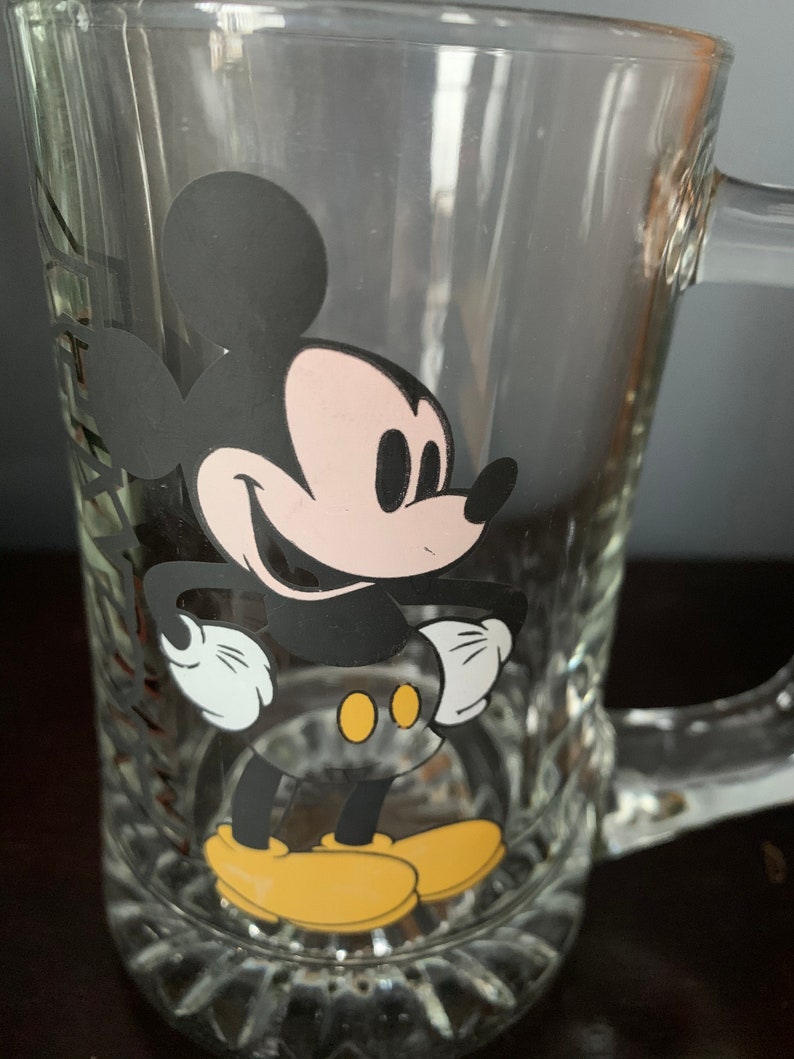 May include: A clear glass mug with a black and white image of Mickey Mouse on the front. Mickey Mouse is smiling and has his hands on his hips. He is wearing yellow shoes.