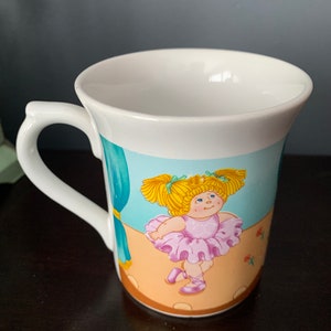 Könnte beinhalten: Weiße Keramiktasse mit einer bunten Illustration einer Cabbage Patch Kid Puppe, die als Ballerina verkleidet ist. Die Puppe trägt einen rosa Tutu und rosa Ballettschuhe. Der Hintergrund ist ein blauer Bühnenvorhang mit einem braunen Bühnenboden.