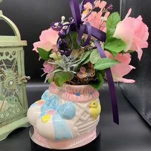 Könnte beinhalten: Eine keramische Ostern-Korb-förmige Vase, weiß mit rosa und blauen Akzenten bemalt, hält rosa Rosen und Grünzeug. Ein lila Band und ein kleines Schild mit der Aufschrift "Frohe Ostern" sind an der Anordnung befestigt. Auf der Seite befindet sich ein gelbes Küken.