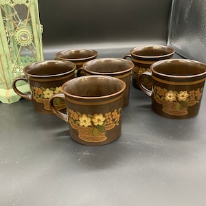 May include: Six brown ceramic mugs with a floral design. The mugs have a brown and gold floral pattern with a basket design.