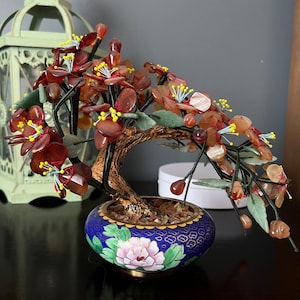 Puede incluir: Un árbol bonsái de piedras preciosas rojas y verdes en una maceta de cloisonné azul y dorado con un diseño floral. El árbol está hecho de pequeñas piedras pulidas y tiene una forma realista.