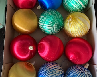 May include: A box of 12 vintage Christmas ornaments. The ornaments are made of a shiny, satin-like material and come in a variety of colors, including red, blue, green, and gold. Some of the ornaments have a striped pattern.