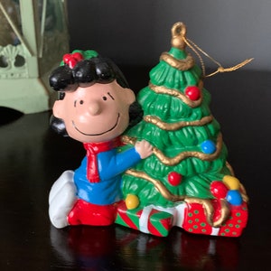May include: A ceramic Christmas ornament featuring a cartoon girl with black hair and a red bow holding a green Christmas tree with gold tinsel and red and blue ornaments. The ornament is decorated with red and green gift boxes.
