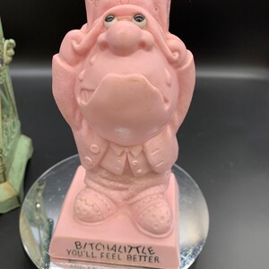 May include: Pink figurine of a man with a sad expression, wearing a hat and a jacket. The figurine is standing on a round base with the text "B/TCHALITTLE YOU'LL FEEL BETTER".