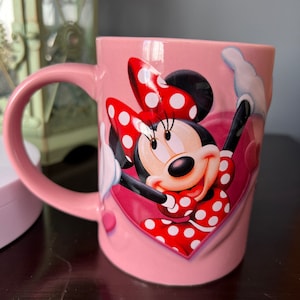 May include: Pink ceramic mug with a graphic of Minnie Mouse wearing a red and white polka dot dress and bow. The mug has a heart-shaped design.