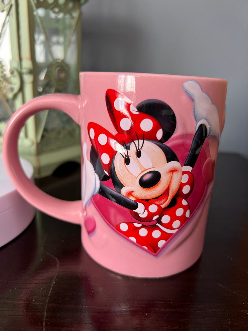May include: Pink ceramic mug with a graphic of Minnie Mouse wearing a red and white polka dot dress and bow. The mug has a heart-shaped design.