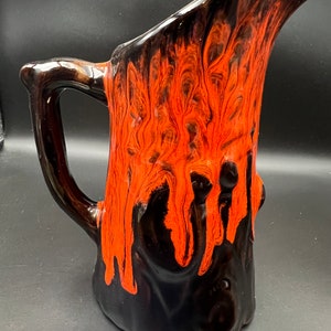 Puede incluir: Una jarra de cerámica negra y naranja con un diseño de tronco de árbol. La jarra tiene una boca ancha y un asa.