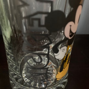 May include: A clear glass beer mug with a black and yellow Mickey Mouse design. The mug has a textured bottom.