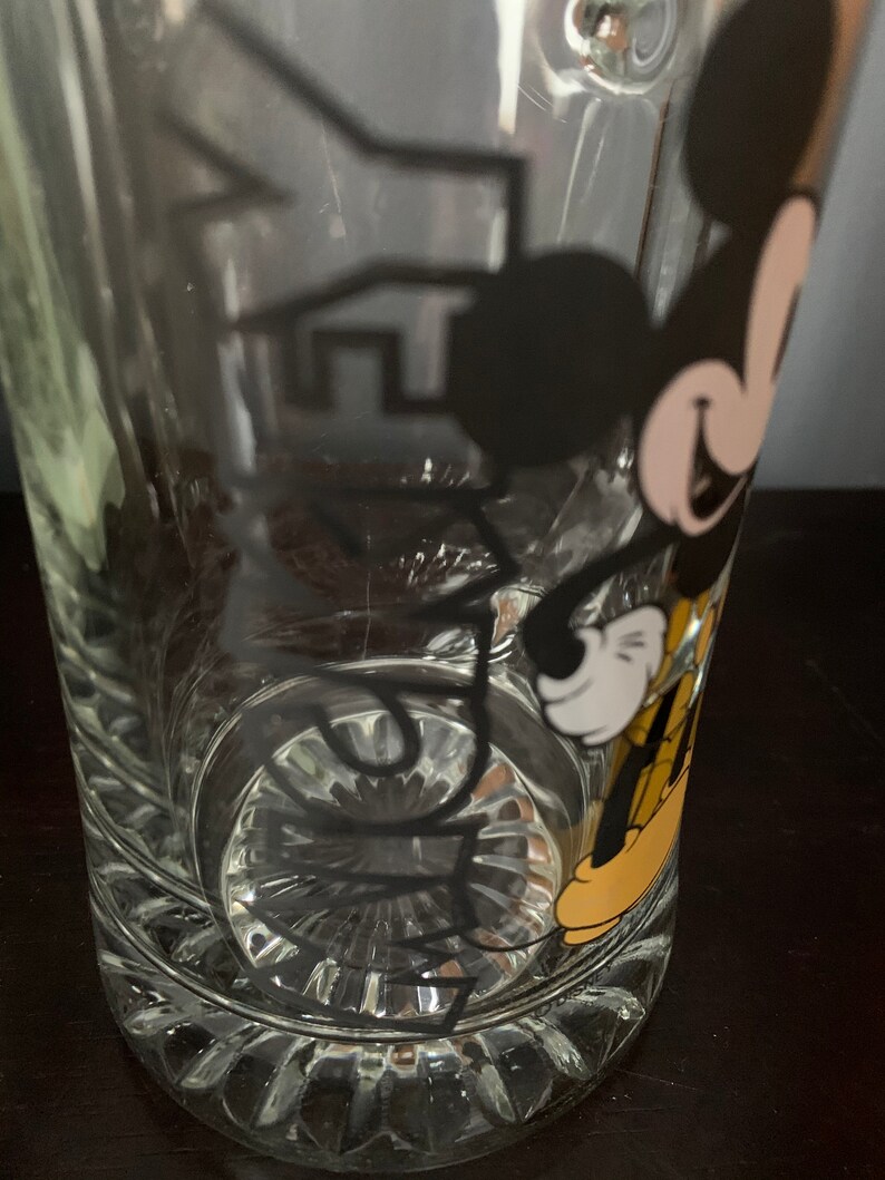 May include: A clear glass beer mug with a black and yellow Mickey Mouse design. The mug has a textured bottom.