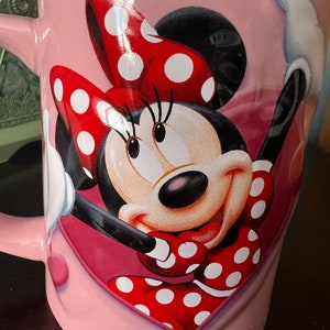 May include: Pink ceramic mug with a red and white polka dot Minnie Mouse design. Minnie Mouse is smiling and has her arms raised. The mug has a heart shape around Minnie Mouse.