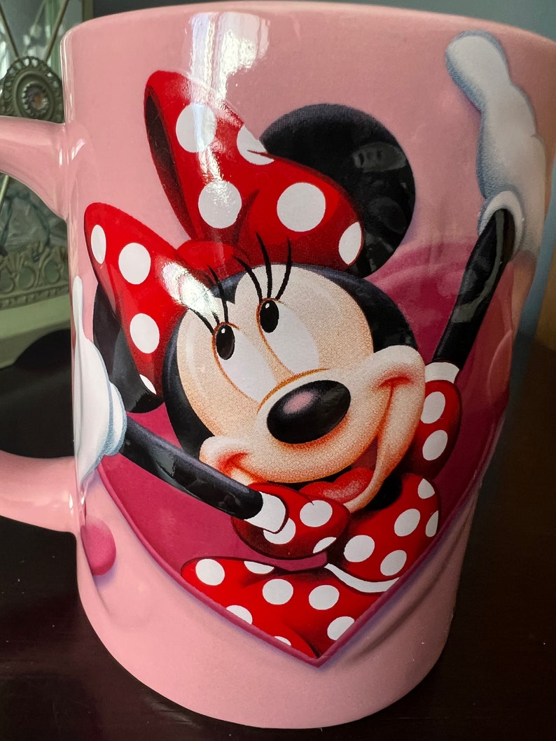 May include: Pink ceramic mug with a red and white polka dot Minnie Mouse design. Minnie Mouse is smiling and has her arms raised. The mug has a heart shape around Minnie Mouse.