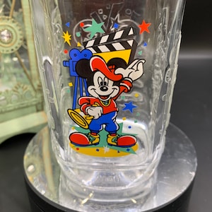 Puede incluir: Un vaso de vidrio transparente con un diseño de Mickey Mouse. Mickey lleva un sombrero rojo y sostiene un micrófono. El vaso tiene un diseño colorido con estrellas, confeti y una claqueta de cine.
