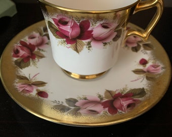 Pode incluir: Uma chávena de chá e um pires brancos com uma borda dourada e um padrão de rosas cor-de-rosa e vermelhas. A chávena tem uma pega dourada.