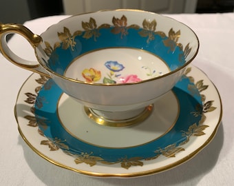 May include: A white teacup and saucer with a teal blue band and gold leaf accents. The teacup features a floral design with pink, yellow, and blue flowers.