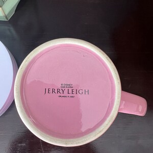 May include: Pink ceramic mug with a white rim and a handle. The bottom of the mug has the text "&copy; DISNEY MADE IN CHINA JERRY LEIGH ORLANDO, FL 32837"