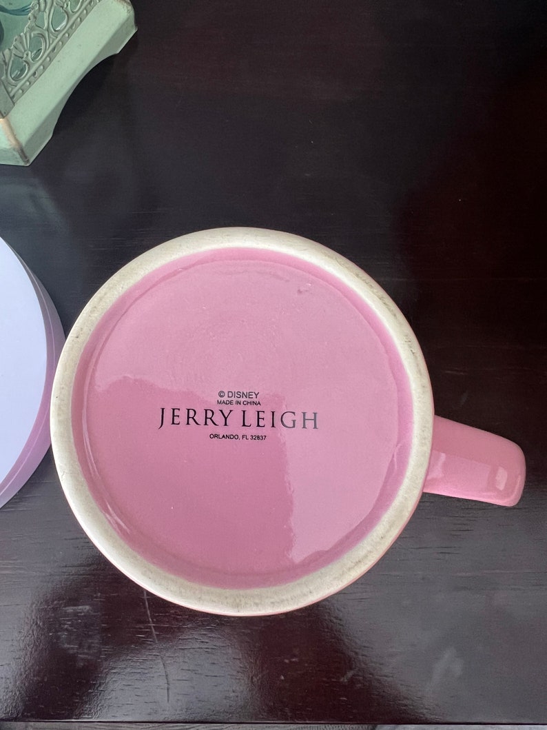 May include: Pink ceramic mug with a white rim and a handle. The bottom of the mug has the text "&copy; DISNEY MADE IN CHINA JERRY LEIGH ORLANDO, FL 32837"