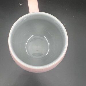 May include: A top-down view of a round mug featuring a soft pink exterior and a smooth, light grey interior. The mug has a single, matching pink handle extending from the side, and the circular opening reveals a deep, clean basin.