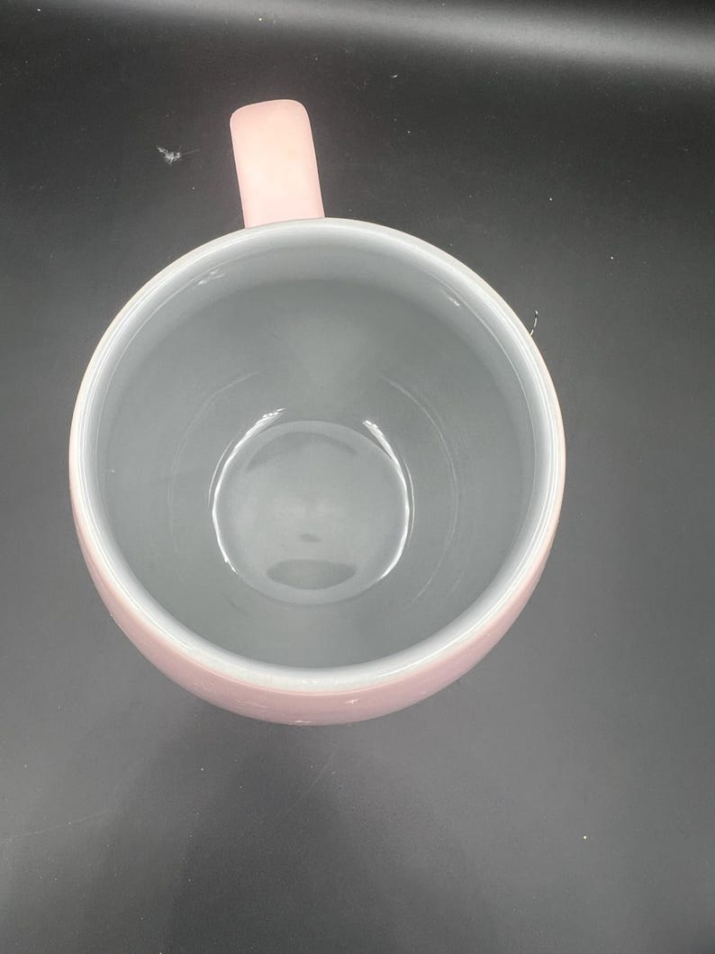 May include: A top-down view of a round mug featuring a soft pink exterior and a smooth, light grey interior. The mug has a single, matching pink handle extending from the side, and the circular opening reveals a deep, clean basin.