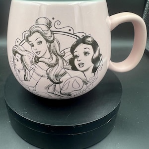 May include: A light pink ceramic mug with a rounded shape and a handle on the right side. The exterior features a black line art illustration of three female characters enclosed within a decorative circular frame on the front of the mug.