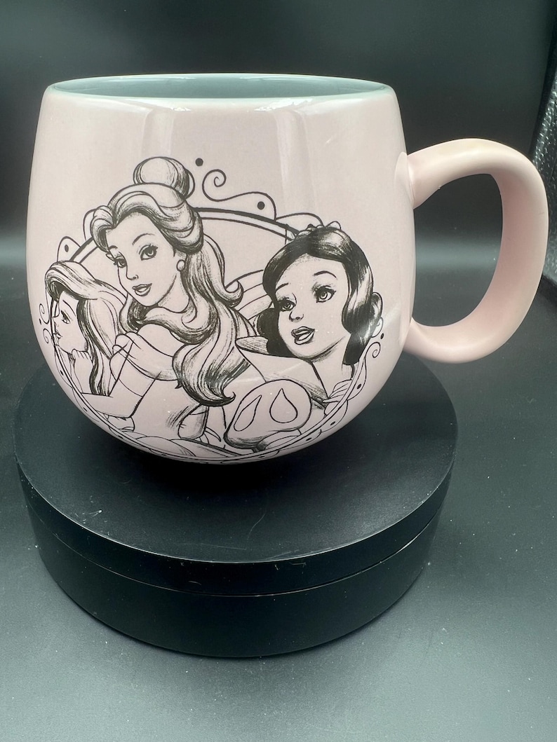 May include: A light pink ceramic mug with a rounded shape and a handle on the right side. The exterior features a black line art illustration of three female characters enclosed within a decorative circular frame on the front of the mug.