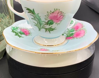 May include: A delicate teacup and saucer set with a light blue background and pink and green floral designs. The set is on a black circular stand.