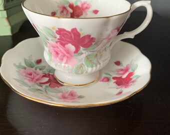 May include: A white teacup and saucer with a floral pattern of pink and red roses. The cup and saucer have a gold trim.