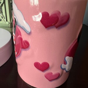 May include: Pink ceramic mug with a cartoon design featuring Mickey Mouse and Minnie Mouse with hearts. The mug has a glossy finish.