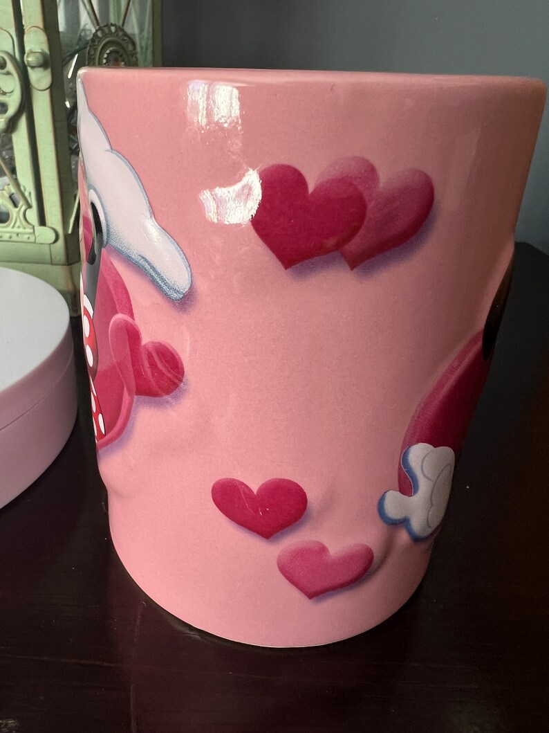 May include: Pink ceramic mug with a cartoon design featuring Mickey Mouse and Minnie Mouse with hearts. The mug has a glossy finish.