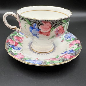 May include: A white teacup and saucer set with a floral pattern. The cup and saucer feature a black and white lattice design with pink, blue, and green flowers.