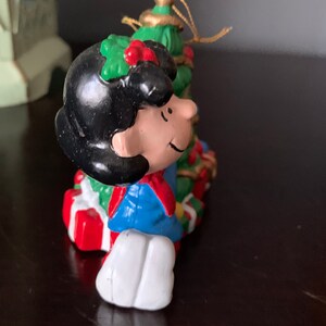 May include: A Christmas ornament featuring a cartoon character with black hair, a blue jacket, and a red scarf, holding a green Christmas tree with red and gold ornaments.