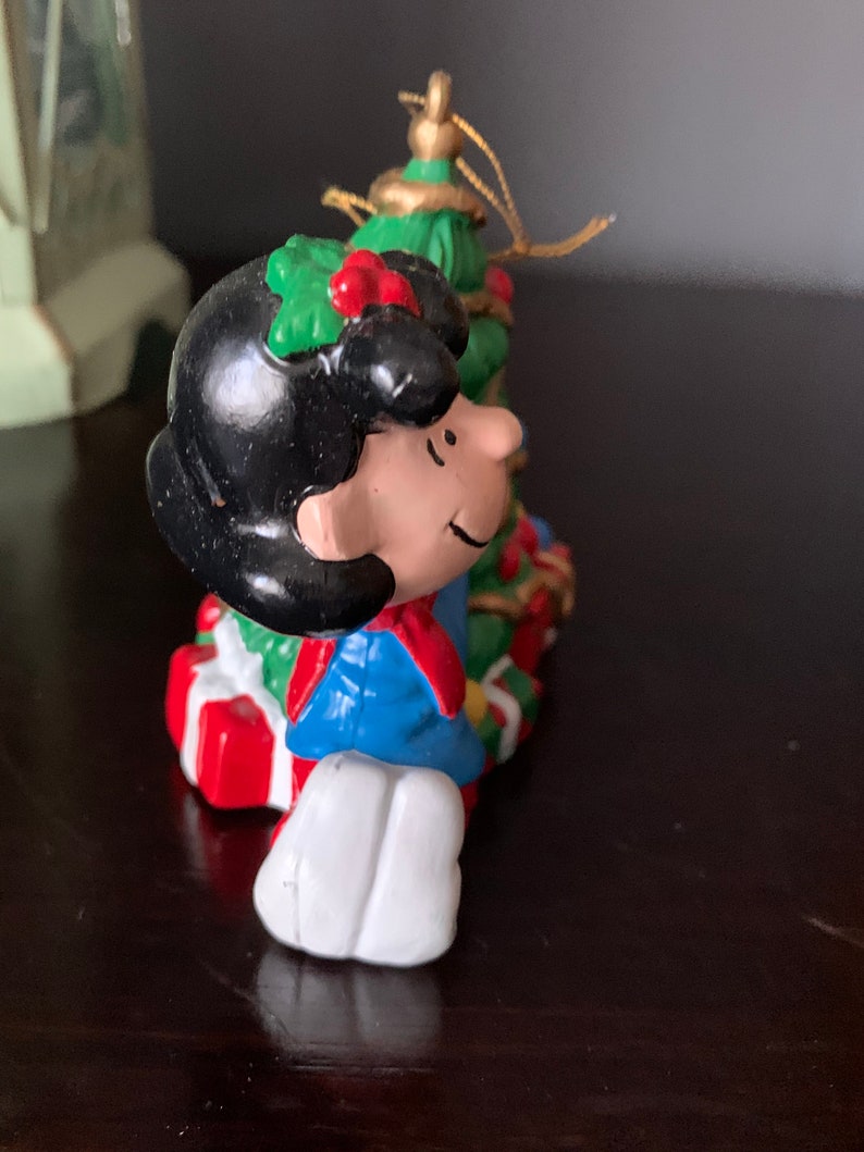 May include: A Christmas ornament featuring a cartoon character with black hair, a blue jacket, and a red scarf, holding a green Christmas tree with red and gold ornaments.